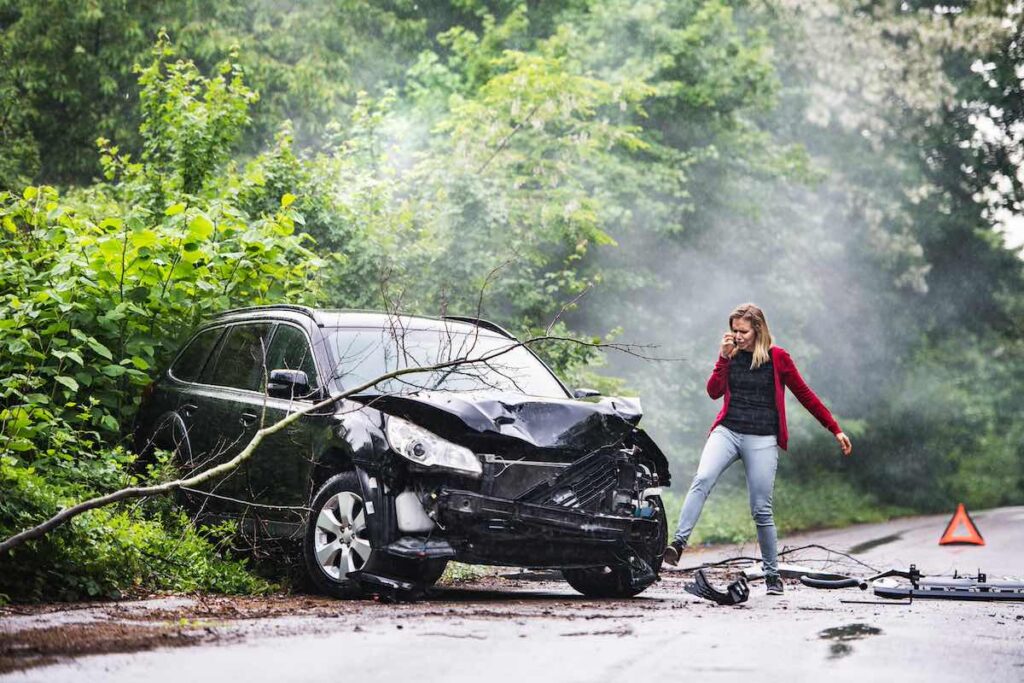An angry and frustrated young woman with a smartphone beside the damaged car after a car accident